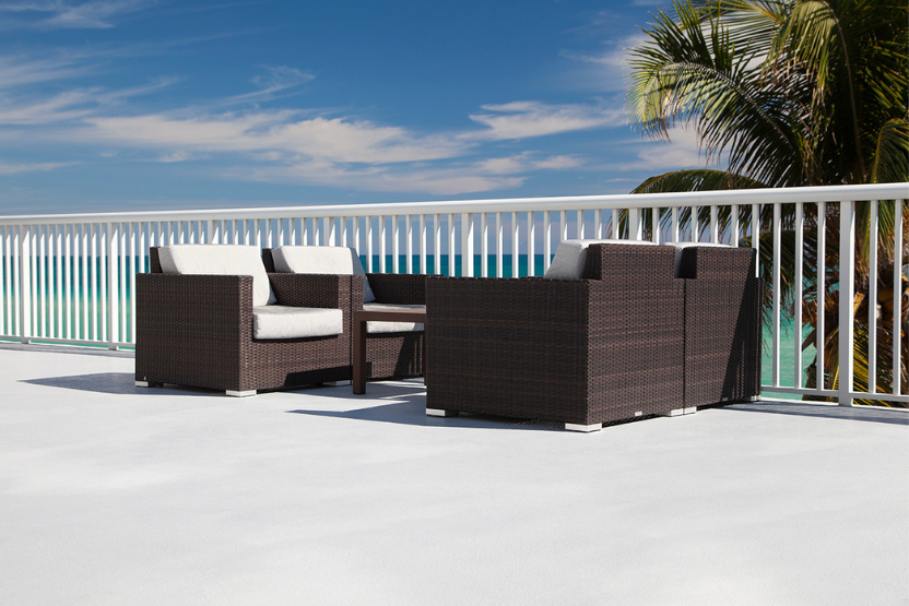 Outdoor wicker lounge chairs with white cushions and a matching coffee table on a patio overlooking the ocean, with palm trees and a white railing in the background.